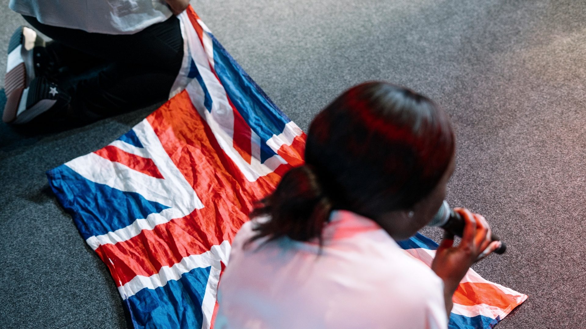 A Union Jack laid on carpet as a young woman kneels to pray over it.